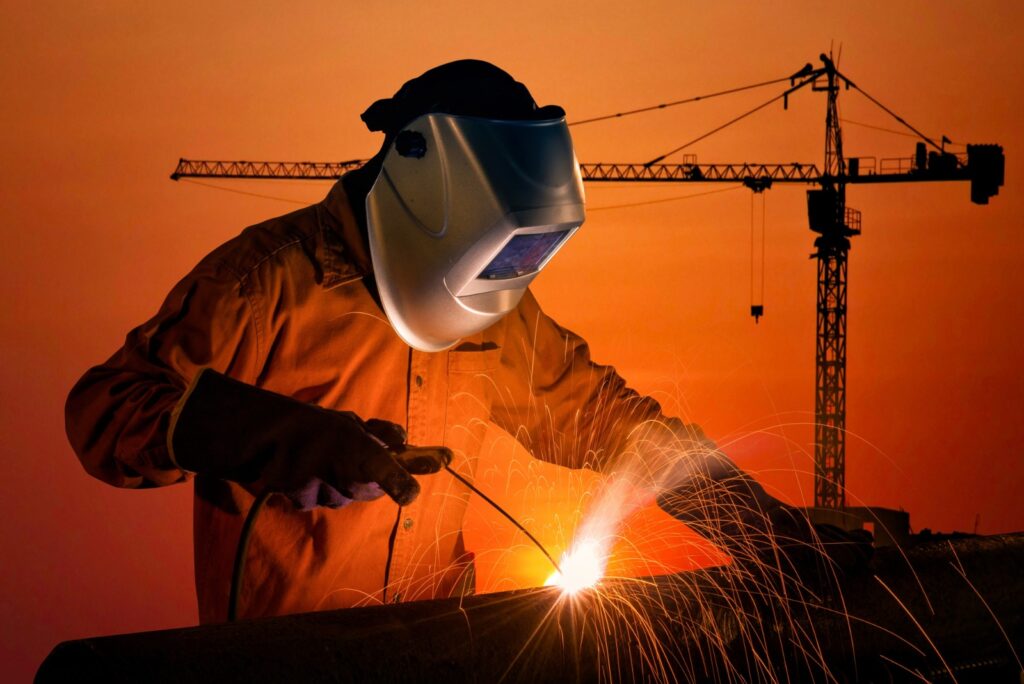 Welder in protective gear working at sparks flying from metal with construction cranes in the background.