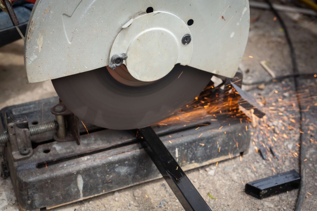 Cutting metal with an abrasive saw, showing sparks flying as the rotating blade slices through a black metal bar on a workshop floor.